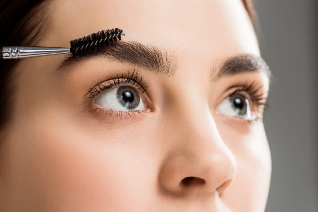 cropped view of young woman styling eyebrow with eyebrow brush on grey
